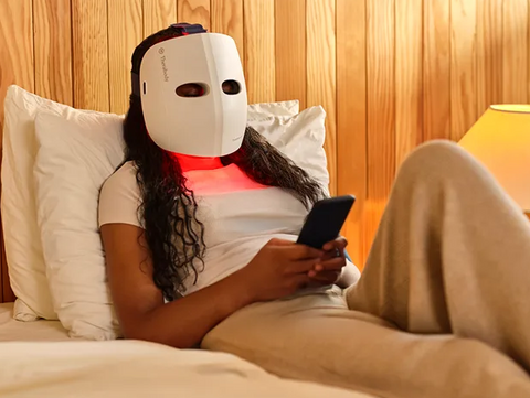 Person lying on a bed wearing a white mask with red lights, using a phone.