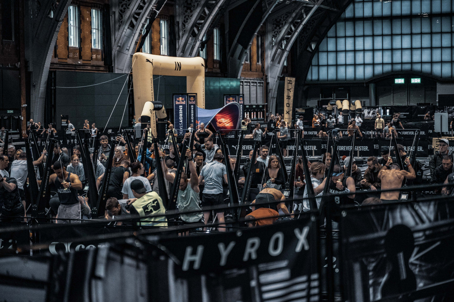 Large indoor event space with a crowd of people, some holding up signs with 'HYROX' branding.
